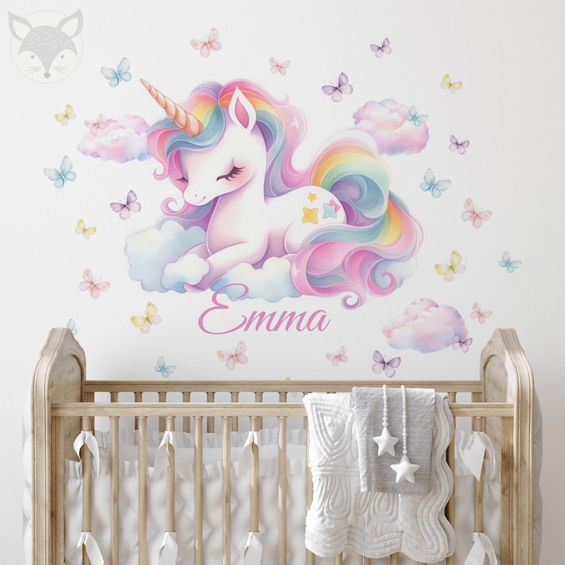 May include: A whimsical wall decal featuring a pastel unicorn with a rainbow mane and tail, resting on fluffy clouds. Butterflies in various colors surround the unicorn. The name "Emma" is written in cursive below the unicorn.