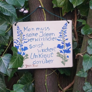 Könnte beinhalten: Ein weißes Keramik Schild mit blauen Blumen und dem Zitat "Man muss seine Ideen verwirklichen sonst wuchert Unkraut darüber" von Jean Paul.