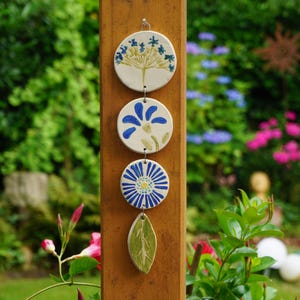 Könnte beinhalten: Ein dekorativer Gartenhänger mit vier Keramikteilen. Die oberen drei sind rund mit blauen Blumenmustern, das untere Teil ist blattförmig und grün. Sie hängen an einem Holzpfosten, mit einem üppigen grünen Garten im Hintergrund.