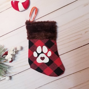 May include: A small red and black plaid stocking with a white paw print and a red heart in the center. The stocking has a brown faux fur cuff and a red and orange loop for hanging.