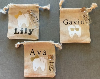 Può includere: Tre piccole borse di tessuto beige con un disegno di dente e una piccola bottiglia di vetro con un tappo di sughero. Ogni borsa ha un nome scritto: Lily, Gavin e Ava.