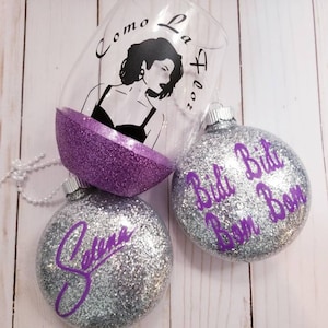 May include: A clear wine glass with a purple glitter base and a black silhouette of a woman, with the words "Como La Flor". Two silver glitter ornaments with purple text that reads "Selena" and "Bidi Bidi Bom Bom".