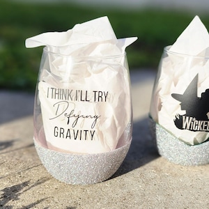 May include: Two silver glitter wine glasses with black vinyl decals. One glass says "I think I'll try defying gravity" and the other says "Wicked" with a silhouette of the Wicked Witch of the West.