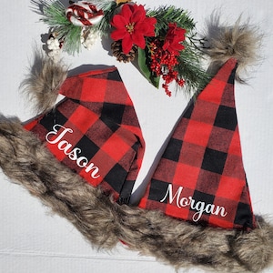 May include: Two red and black buffalo plaid Santa hats with faux fur trim. The hats are personalized with the names "Jason" and "Morgan" in white lettering.