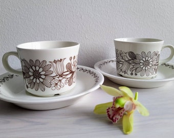 May include: Two white teacups with brown floral designs and matching saucers. The cups have a simple, elegant design with a brown floral pattern around the rim. The saucers have a matching floral pattern around the edge.