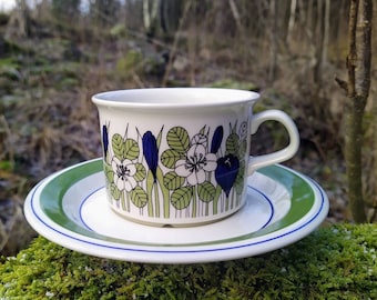 May include: A white ceramic coffee cup and saucer set with a floral design. The cup features a pattern of white flowers, green leaves, and blue accents. The saucer has a green and blue rim. The set is placed on a bed of green moss.