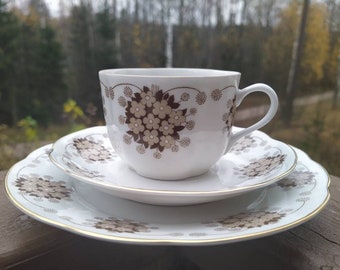 May include: A white teacup and saucer set with a floral design in brown and tan. The set includes a teacup, a small saucer, and a larger plate, all with gold trim. The background is a blurred view of trees.
