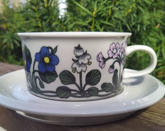 May include: A white porcelain teacup and saucer with a floral design. The design features blue, green, and pink flowers.