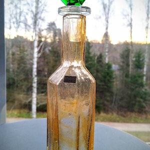 May include: A clear glass bottle with a textured surface and a green glass butterfly stopper. The bottle has a label that says "ARABIA".