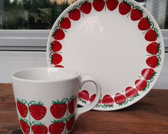 May include: A white ceramic mug with a strawberry pattern and a white ceramic plate with a strawberry pattern.