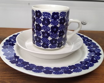 May include: A white ceramic mug, bowl, and plate set. The mug and plate have a blue floral pattern. The bowl is plain white. The mug is sitting on the bowl, which is on the plate. The set is on a wooden surface.