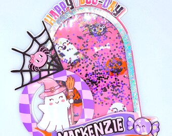 Op de afbeelding: Een decoratieve Halloween-taarttopper met een roze boog gevuld met glitters, een geest met een heksenhoed, een spin in een web en de tekst HAPPY BOO-DAY en MACKENZIE. Het ontwerp bevat paarse en witte geblokte accenten.