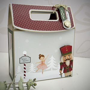 Peut inclure: Une boîte cadeau de Noël décorative avec un toit à carreaux rouges et blancs et une poignée. La boîte présente une scène hivernale avec une ballerine, un casse-noisette et un panneau indiquant "Polo Norte". Une étiquette avec le mot "JOY" est attachée.