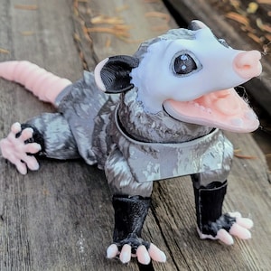 May include: A detailed, 3D-printed opossum figurine. The opossum has a white face, pink nose and mouth, and gray fur. Its tail is pink, and it has black paws. The figurine is posed on a wooden surface.