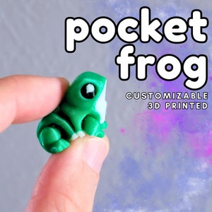 May include: A small, green 3D-printed frog figurine with white accents, held between two fingers. The text "pocket frog" is above the frog, and "CUSTOMIZABLE 3D PRINTED" is below. The background is a gradient of purple and gray.