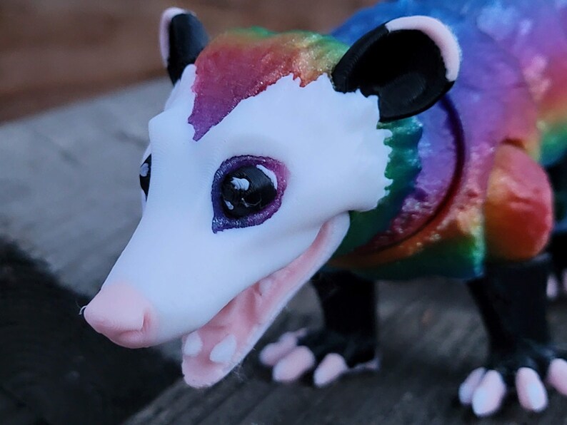 May include: A colorful figurine of an opossum with a rainbow-colored back and head. The opossum has a white face, pink nose, and open mouth, revealing small teeth. Its ears and paws are black, and the eyes are dark with purple accents.