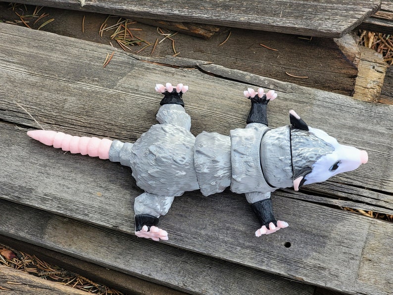 May include: A gray articulated opossum figurine with a pink tail and feet, black paws, and a white face. The toy is lying on a weathered wooden surface.