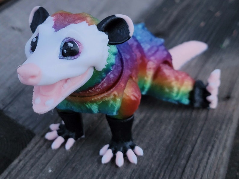 May include: A colorful opossum figurine with a rainbow-colored body, white face, and pink mouth and paws. The figurine has black ears and feet, and is posed in a playful stance. The figurine is a unique decorative item.