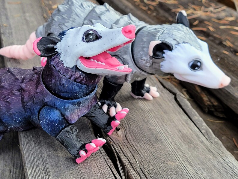 May include: Two detailed opossum figurines. One has a dark purple and blue body with a white face and pink mouth and feet. The other is gray with a white face and pink nose and feet. Both are posed on a wooden surface.