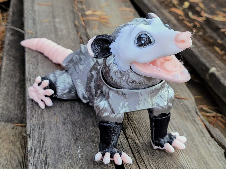 May include: A detailed, 3D-printed opossum figurine. The opossum has a white face, pink nose and mouth, and gray fur. Its tail is pink, and it has black paws. The figurine is posed on a wooden surface.