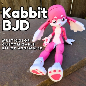 May include: A pink and white articulated doll in the shape of a rabbit. The doll has large ears, a white face, and pink paws. The text "Kabbit BJD" and "MULTICOLOR CUSTOMIZABLE KIT OR ASSEMBLED" are displayed.