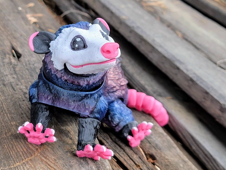 May include: A whimsical figurine of an opossum with a white face, pink nose, and pink feet. The opossum's body is a mix of blue, purple, and black, with a starry effect. The ears are black with pink trim, and the tail is pink.