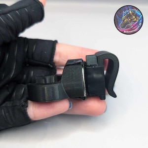 Puede incluir: Se muestra un dispositivo negro impreso en 3D, posiblemente un accesorio para guantes. El dispositivo tiene un asa curva y un cuerpo rectangular. El dispositivo está sujeto a un guante negro. Una pegatina circular con una criatura con forma de caballo está en el fondo.