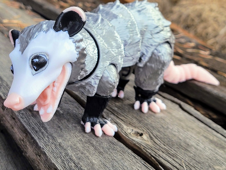 May include: A 3D-printed opossum figurine with a detailed design. The opossum has a white face, pink mouth and paws, and a gray body with a segmented, armored appearance. Its tail is pink, and it stands on a wooden surface.