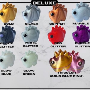 May include: A collection of Deluxe figurines in various colors, including gold, silver, copper, and marble. The figurines have a dinosaur-like design with the text "DELUXE" at the top and color labels like "BLACK GLITTER" and "GLOW BLUE".