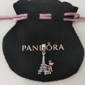 May include: A silver charm with a red heart and Eiffel Tower design hangs from a black pouch with the word "PANDORA" printed on it. The charm also features a small figure with a red hat and scarf.