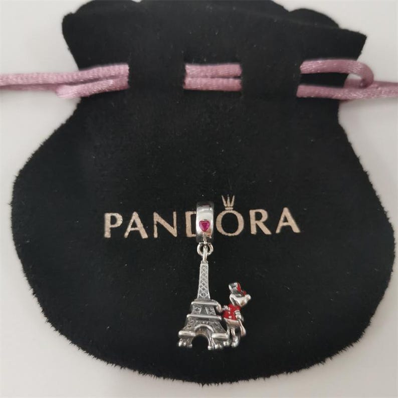 May include: A silver charm with a red heart and Eiffel Tower design hangs from a black pouch with the word "PANDORA" printed on it. The charm also features a small figure with a red hat and scarf.