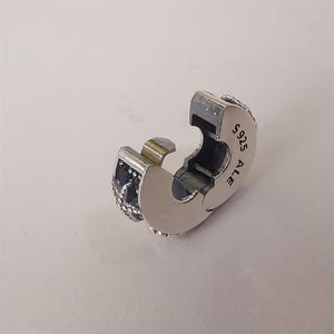 Pode incluir: Um charm prateado com um design articulado. O charm apresenta uma seção decorativa com detalhes em azul escuro e pequenas pedras transparentes. O texto "S925 ALE" é visível na lateral do charm.