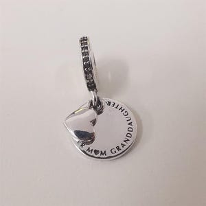 May include: A silver charm pendant with a heart and a round tag. The tag is inscribed with the words "GRANDDAUGHTER" and "MOM". The charm has a small loop with black accents.