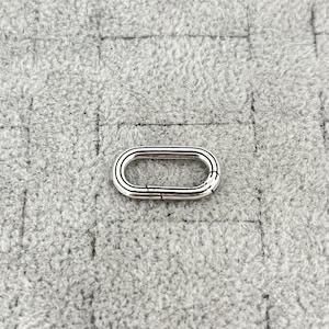 Op de afbeelding: Een zilverkleurige ovale sluiting met een scharnierend ontwerp voor een veilige sluiting. De sluiting is geplaatst tegen een getextureerde grijze achtergrond, die de metallic glans en ingewikkelde details benadrukt. Ideaal voor het maken van sieraden of knutselen.