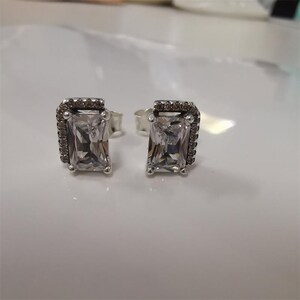 Peut inclure: Une paire de boucles d'oreilles à clous en argent. Chaque boucle d'oreille présente une pierre précieuse rectangulaire et transparente entourée d'un halo de petites pierres sombres. Les boucles d'oreilles sont posées sur une surface blanche.
