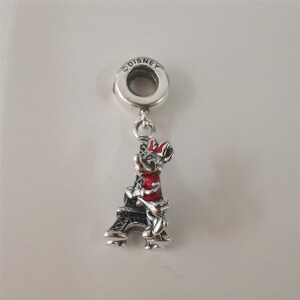 May include: A silver charm featuring Minnie Mouse in a red dress and bow, embracing a black Eiffel Tower. The charm is marked with the Disney logo and is designed to be added to a bracelet or necklace.