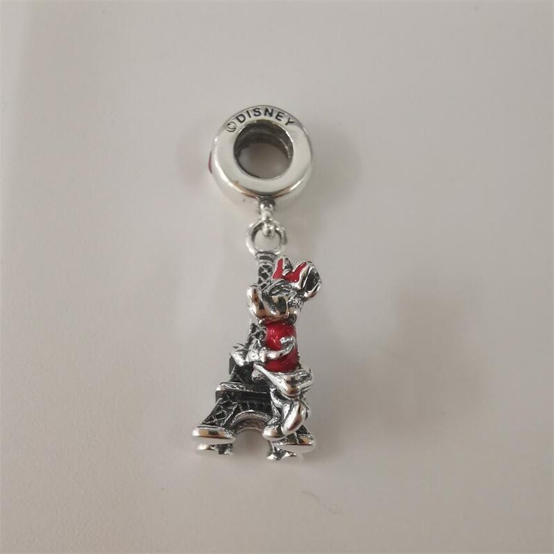 May include: A silver charm featuring Minnie Mouse in a red dress and bow, embracing a black Eiffel Tower. The charm is marked with the Disney logo and is designed to be added to a bracelet or necklace.
