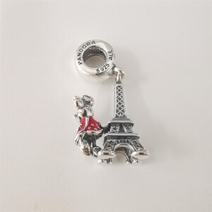 May include: A silver charm featuring a miniature Eiffel Tower and a figure in a red dress. The charm has a loop for attaching to a bracelet or necklace. The charm is marked with "S925" and "ALE".