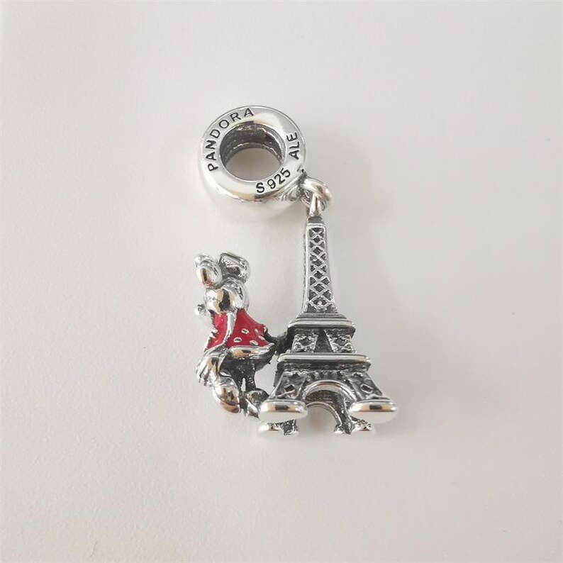 May include: A silver charm featuring a miniature Eiffel Tower and a figure in a red dress. The charm has a loop for attaching to a bracelet or necklace. The charm is marked with "S925" and "ALE".