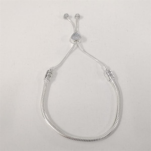 Könnte beinhalten: Ein silberfarbenes Armband mit Schlangenmuster. Das Armband hat einen herzförmigen Anhänger mit kleinen, klaren Steinen. Zwei kleine, runde Charms befinden sich oben am verstellbaren Schieber.