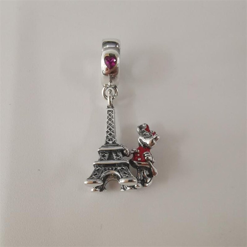 May include: A silver charm featuring the Eiffel Tower and a cartoon character in a red coat and hat. The charm has a heart-shaped gem and is designed to be attached to a bracelet or necklace. The charm is a travel-themed souvenir.