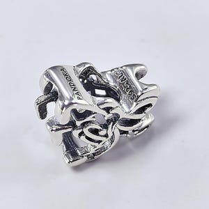 May include: A silver charm with the word "PANDORA" and "S925 ALE" engraved on it. The charm has a heart-shaped design with intricate cutouts and a polished finish. The charm is likely made of sterling silver.