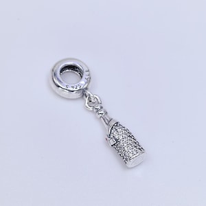 Puede incluir: Un charm de plata con forma de botella de champán. La botella está cubierta de pequeñas piedras brillantes. El charm tiene un lazo en la parte superior para sujetarlo a una pulsera o collar. El charm está marcado con "S925" y "ALE".