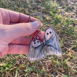 May include: A clear acrylic keyring featuring two cartoon ghosts. One ghost wears a pink bow. The ghosts are grey with black eyes and are attached to a silver heart-shaped keyring. The background is blurred green grass.