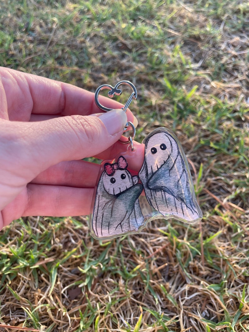 May include: A clear acrylic keyring featuring two cartoon ghosts. One ghost wears a pink bow. The ghosts are grey with black eyes and are attached to a silver heart-shaped keyring. The background is blurred green grass.