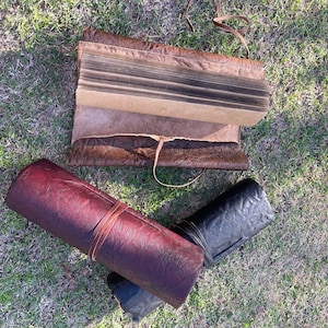 May include: Three rolled-up leather journals with different coloured covers. The journal on the left is a deep burgundy, the one in the middle is brown, and the one on the right is black. All three journals have a leather strap closure.