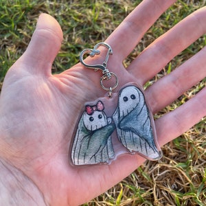 May include: A clear acrylic keyring featuring two cartoon ghosts. One ghost has a pink bow, and both have black eyes. The keyring has a silver heart-shaped clasp.