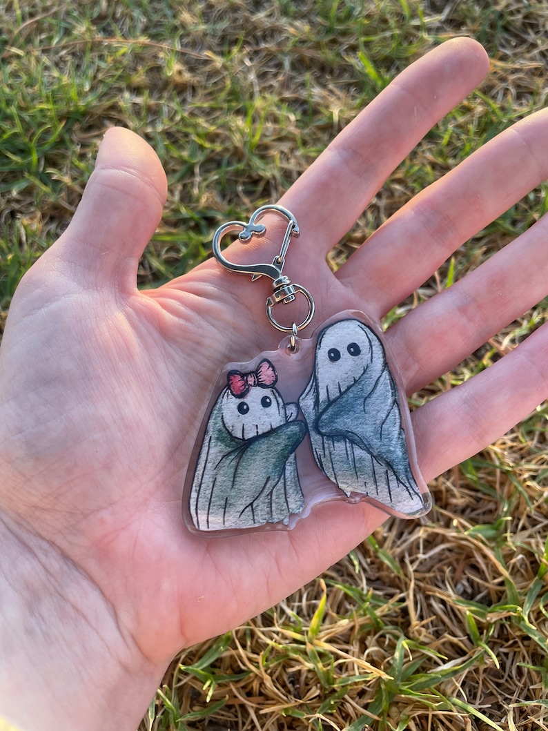 May include: A clear acrylic keyring featuring two cartoon ghosts. One ghost has a pink bow, and both have black eyes. The keyring has a silver heart-shaped clasp.