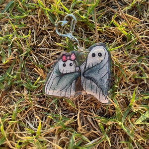 May include: A clear acrylic keyring featuring two cartoon ghosts. One ghost has a pink bow. The ghosts are wrapped in grey sheets with black eyes. The keyring has a silver heart-shaped clasp, set against a backdrop of dry grass.