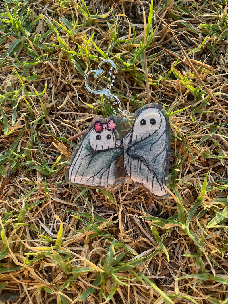 May include: A clear acrylic keyring featuring two cartoon ghosts. One ghost has a pink bow. The ghosts are wrapped in grey sheets with black eyes. The keyring has a silver heart-shaped clasp, set against a backdrop of dry grass.